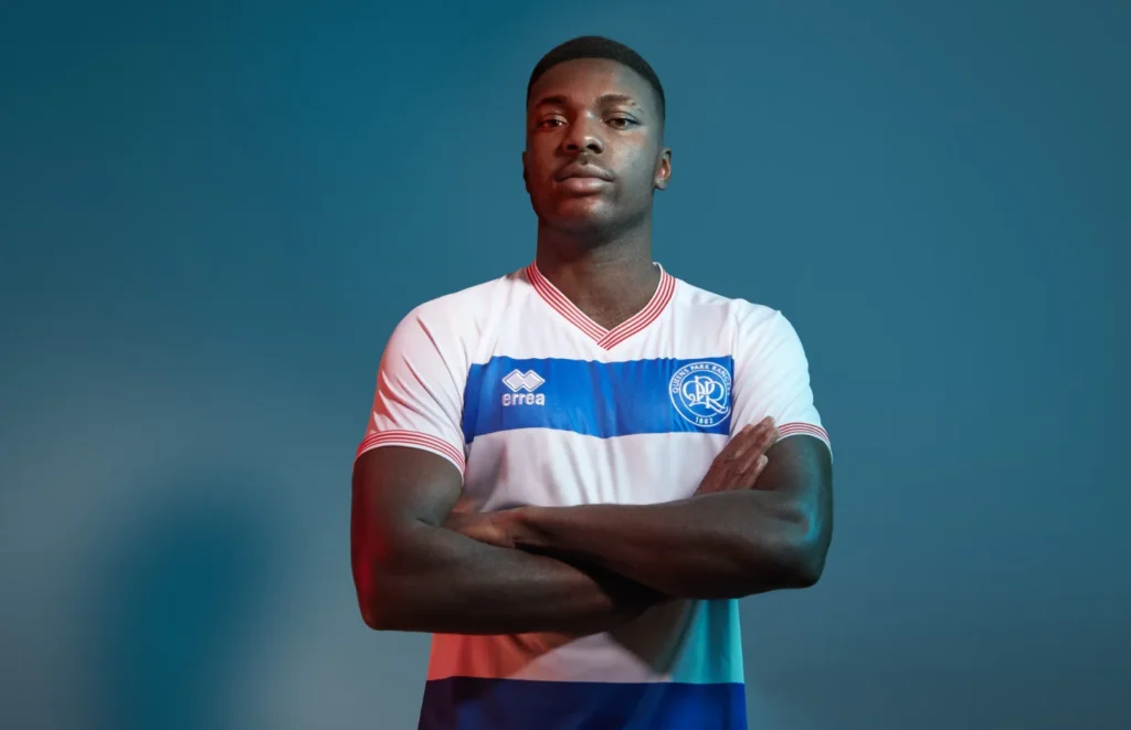 A male soccer player stands with arms crossed, wearing a white and blue striped Queens Park Rangers (QPR) jersey featuring the Kiyan Prince Foundation logo, looking confidently at the camera against a blue background. Long live the prince. Work by MHP Group.