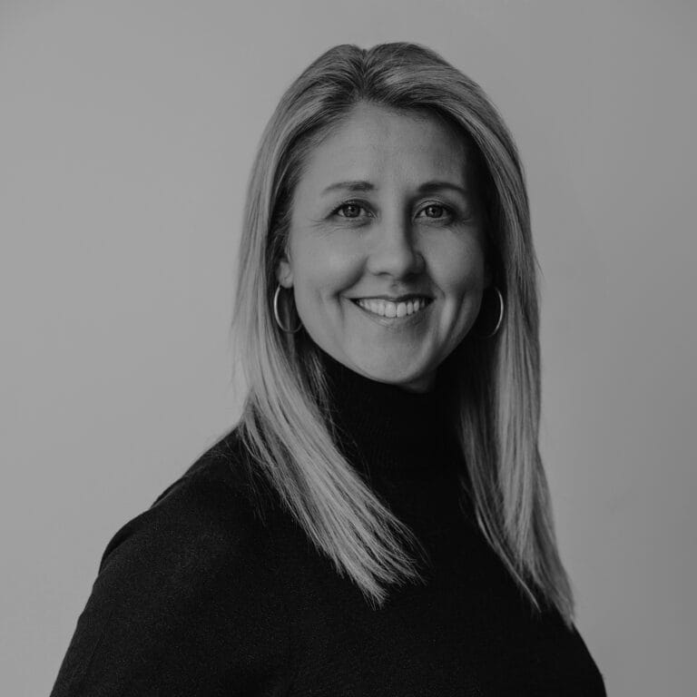 Black and white portrait of Rachel Bower, a woman with straight, shoulder-length hair, wearing hoop earrings and a long-sleeve turtleneck sweater, smiling while looking slightly off camera. Plain background.