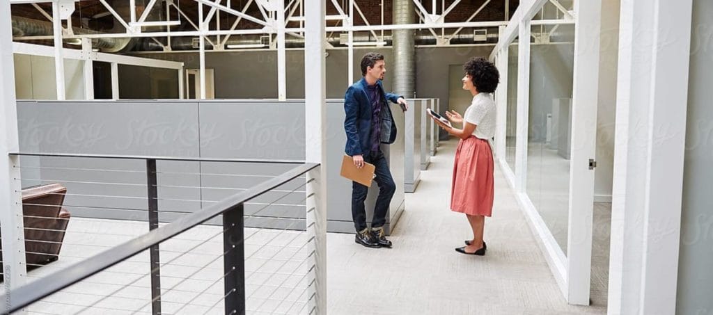 A man and a woman stand in a modern office hallway, having a conversation. The man holds a folder, while the woman gestures with a notepad. The space features white beams, glass walls, and industrial decor.