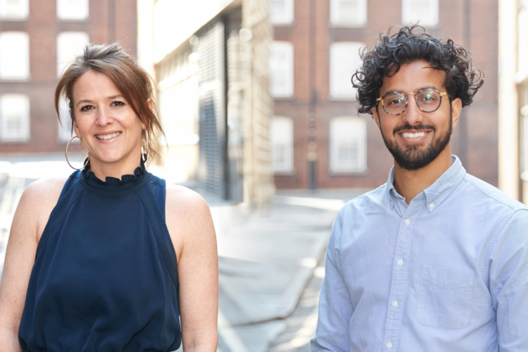 Two people smiling and standing outdoors on a city street with brick buildings in the background, a woman in a navy sleeveless top on the left and a man in glasses and a light blue shirt on the right, discussing healthcare communications.