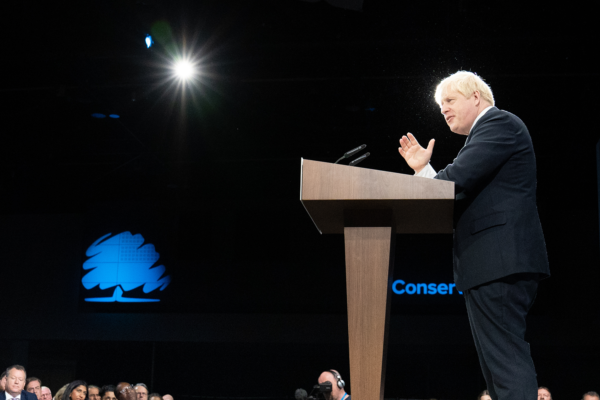 A man in a suit speaks at a podium on stage under a spotlight, with the Conservative Party Conference’s blue tree logo projected behind him. The audience watches attentively from the front.