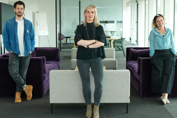 Three members of the Studio Team pose confidently in a modern office lounge—a man in a denim jacket on the left, a woman in black at center, and a woman in a light shirt on the right—showcasing their creative strategy.