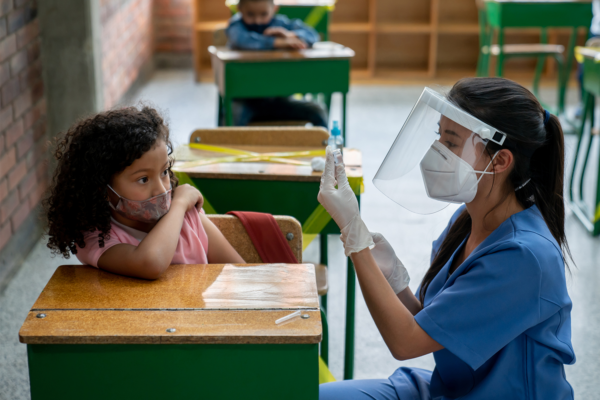 A nurse in scrubs and face shield prepares a syringe while sitting next to a masked young girl at a school desk, reflecting global health efforts in 2022. The girl looks at the nurse, appearing hesitant, as another child sits at a desk in the background.