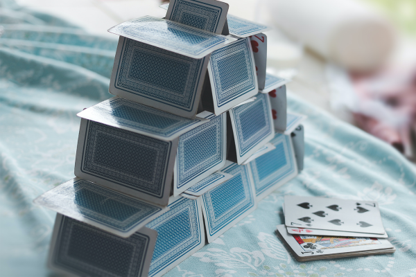 A pyramid-shaped House of Cards sits on a blue patterned cloth, with a small stack of playing cards, including the four of spades and queen of clubs, lying nearby.