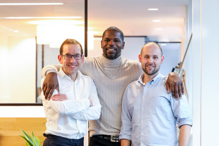 Three men stand closely together in an office setting, smiling at the camera. The man in the middle has his arms around the other two, reflecting a friendly and relaxed atmosphere—highlighting their strategic partnership with Kiyan Prince Foundation.
