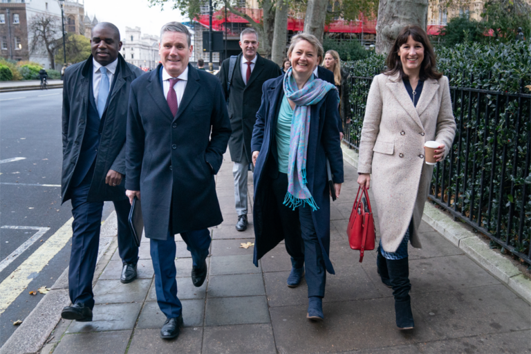 Five people in business attire walk together on a city sidewalk, resembling a group of Political Insider colleagues discussing a recent Labour Reshuffle as they smile and chat past trees and a black fence.