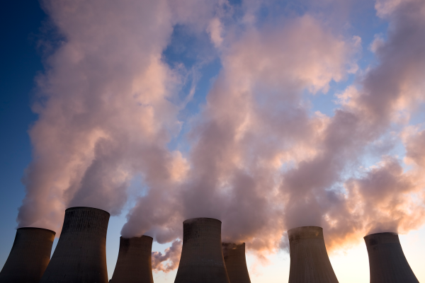 Cooling towers of a power plant emit large plumes of steam or smoke against a blue sky at sunrise, their silhouettes creating a dramatic scene that highlights industrial activity and raises questions about climate change.