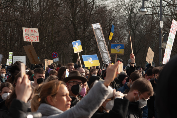 A large crowd of people at a protest, many holding signs with blue and yellow dove designs and Ukrainian flags. Some signs denounce lies or feature photos. Most people are wearing masks, with leafless trees visible in the background.