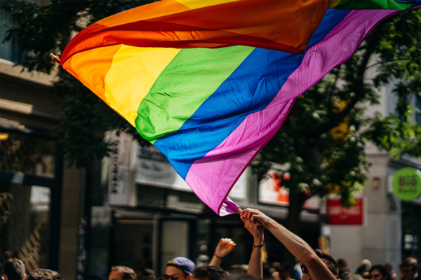 A large rainbow pride flag is being waved by people at an outdoor event during Pride Month, with several hands holding the flagpole and city buildings visible in the background, celebrating visibility and representation.