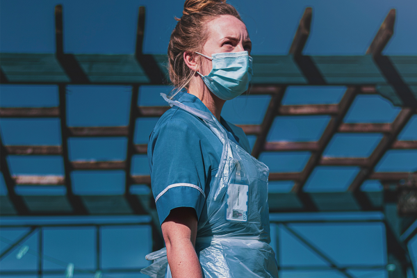 A healthcare worker wearing a blue uniform, plastic apron, and face mask stands outdoors under a wooden pergola, reflecting on COVID-19 recovery beneath a clear blue sky.