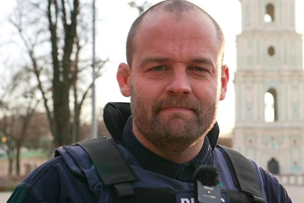 A man with a beard, wearing a bulletproof vest and a microphone, stands outdoors in front of a blurred, ornate tower on a clear day—depicting the BBC Kyiv correspondent reporting from the frontline.