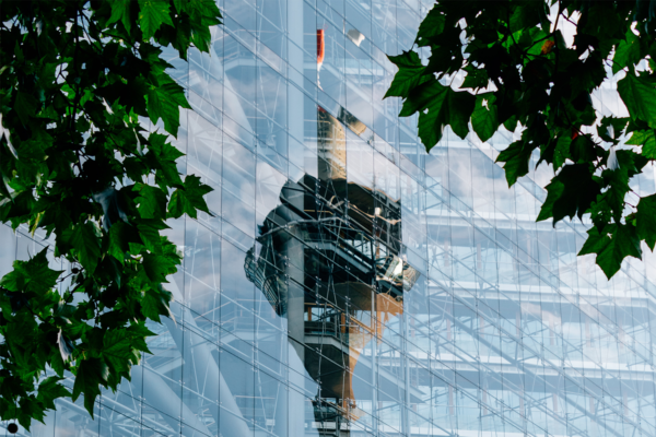 A tower is reflected in the glass windows of a building, with green tree leaves framing the image in the foreground, highlighting a vision of sustainability.