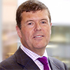 A man in a suit and tie, recognized as a Political Insider, is smiling at the camera with a blurred indoor background. MHP Group discusses social care reforms in their latest edition of Political Insider.