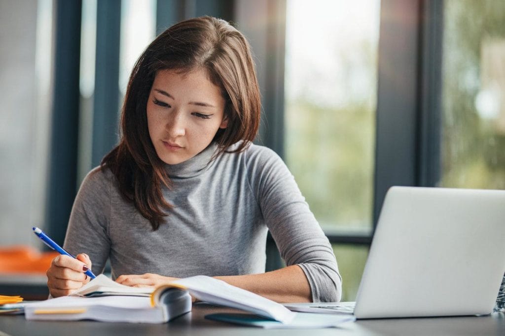A young woman with long brown hair sits at a desk, writing in a notebook with a pen. She is surrounded by open books and has a laptop in front of her, studying in a bright room with large windows.