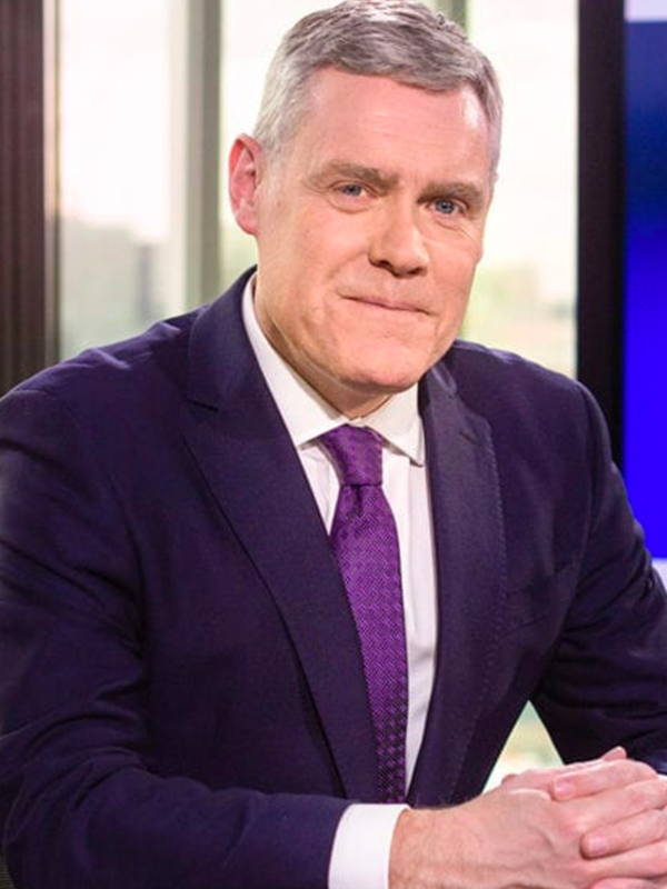 A man with short gray hair in a navy suit, white shirt, and purple tie—resembling top CEOs featured on Sky News—sits at a desk with his hands folded, looking at the camera. Large windows and an out-of-focus cityscape are visible in the background.