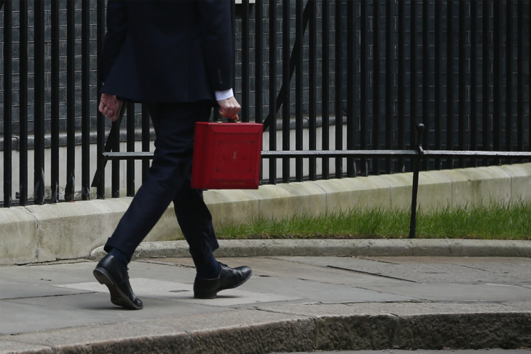 A person in a suit walks on a sidewalk carrying a bright red briefcase, as if preparing for a Cabinet Reshuffle, with a black iron fence and grass in the background. Only the lower half of their body is visible.