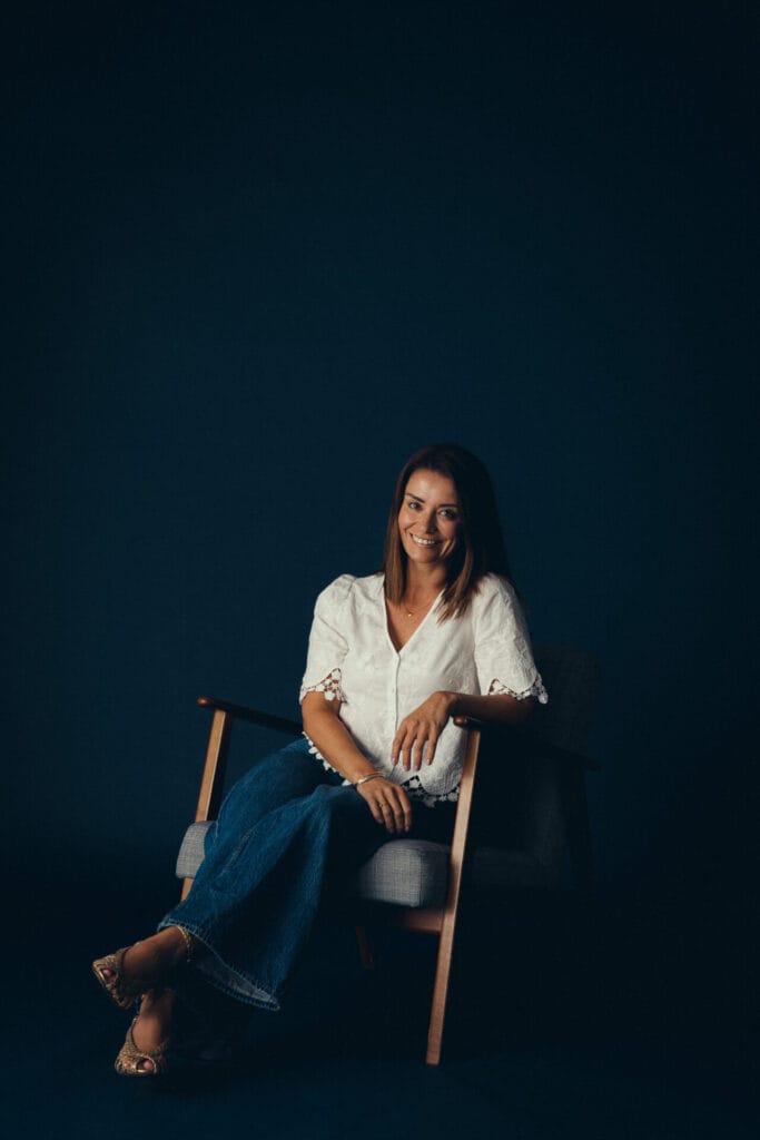 Vicky Purnell, a woman with straight brown hair, wearing a white blouse and blue jeans, sits on a wooden armchair against a dark background, smiling at the camera.