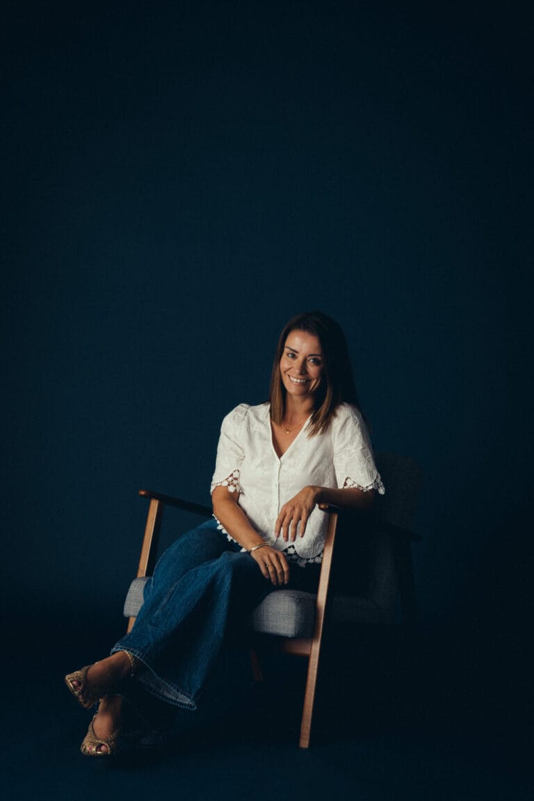 Vicky Purnell, a woman with straight brown hair, wearing a white blouse and blue jeans, sits on a wooden armchair against a dark background, smiling at the camera.