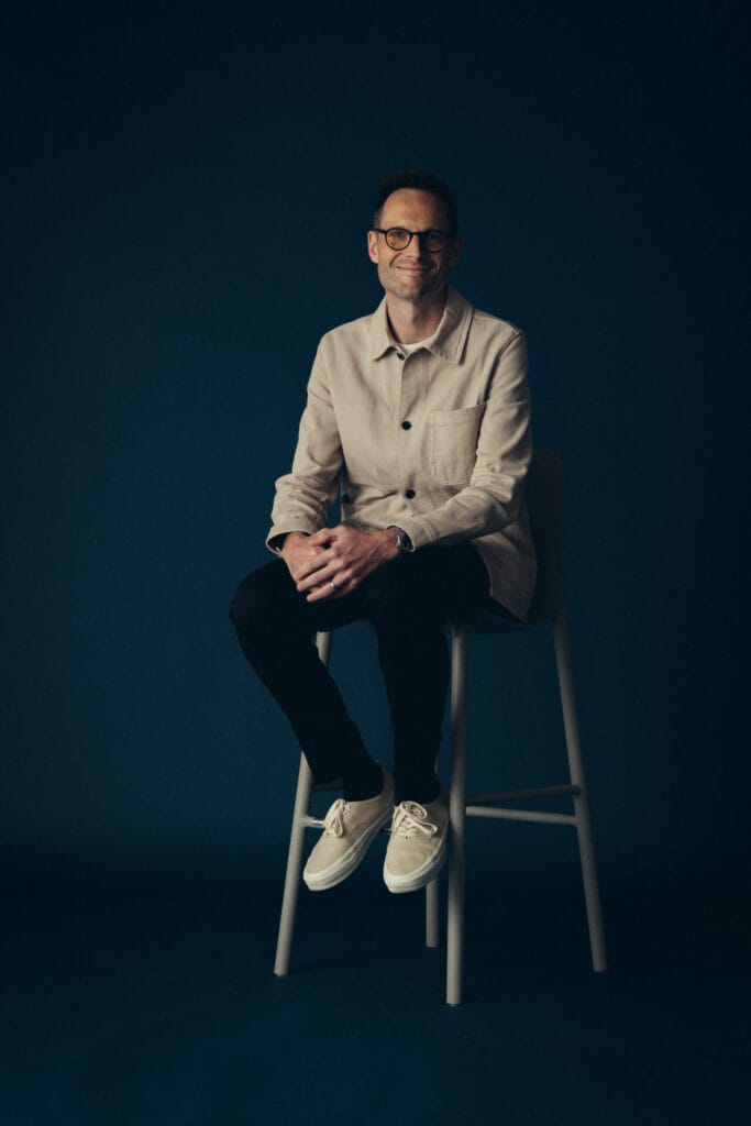 A man wearing glasses, a beige jacket, black pants, and white shoes sits on a tall stool against a dark blue background, smiling with his hands resting on his leg—a stylish look reminiscent of Vicky Purnells signature portrait style.