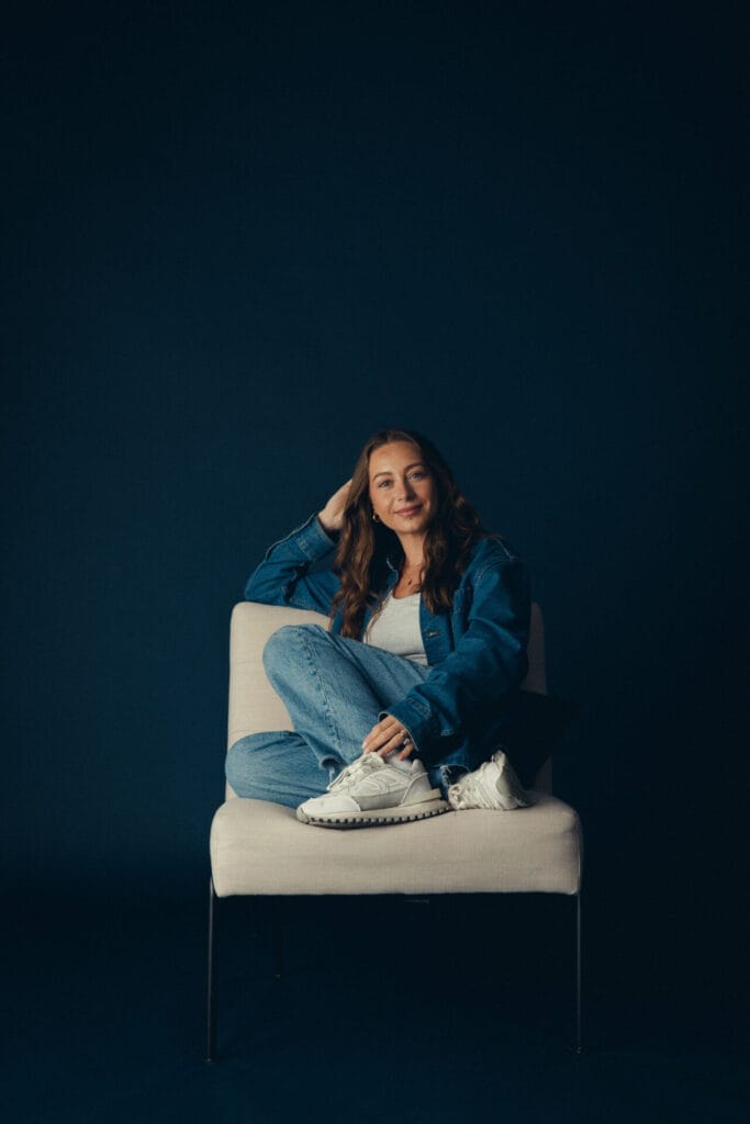 Vicky Purnell, with long brown hair, sits casually on a white chair against a dark blue background, wearing a blue denim jacket, light blue jeans, and white sneakers, one leg crossed over the other and a relaxed expression.