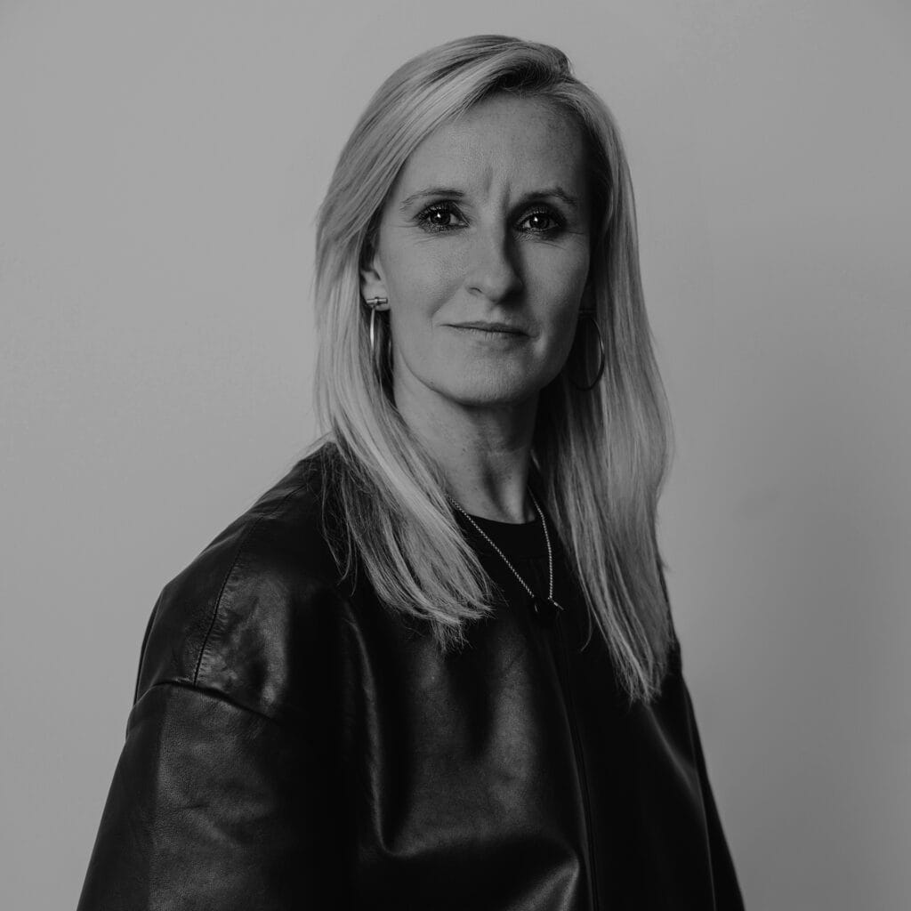 Black and white portrait of Gemma Sawyer, a woman with straight, light-colored hair, wearing a dark jacket and hoop earrings, looking confidently at the camera against a plain background.