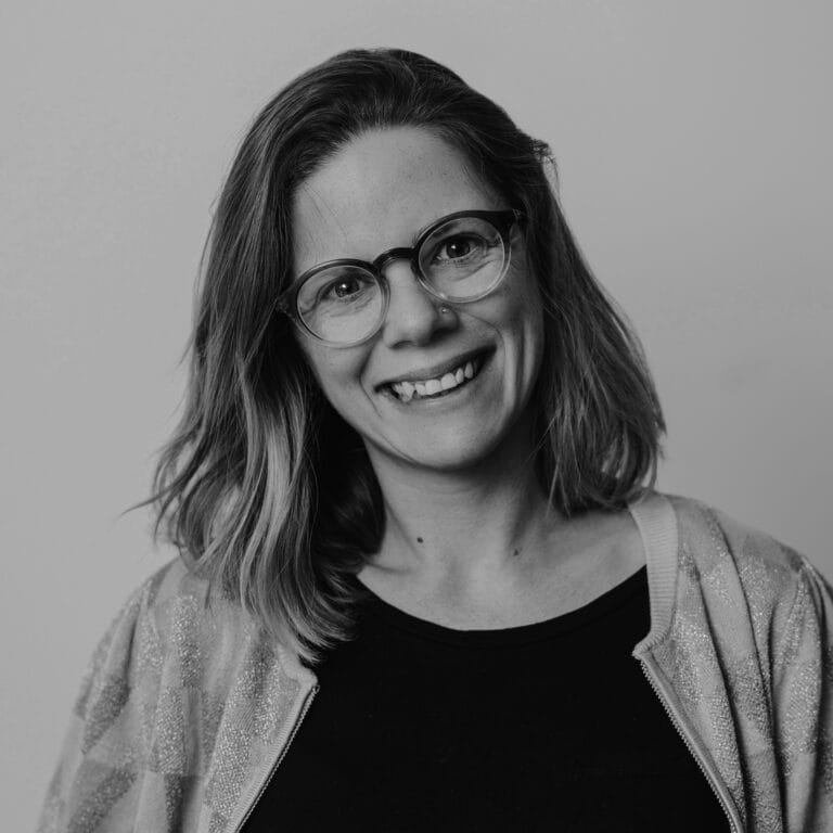 A woman with shoulder-length hair and glasses smiles at the camera. She is wearing a light jacket over a dark top. The photo is in black and white with a plain background.