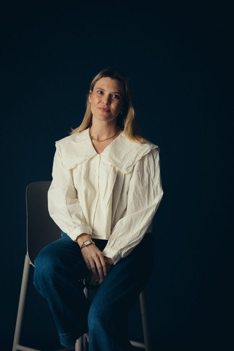 Vicky Purnell, with long blonde hair, sits on a stool against a dark background, wearing a white blouse with a large collar and blue jeans, looking slightly to the side with a gentle expression.