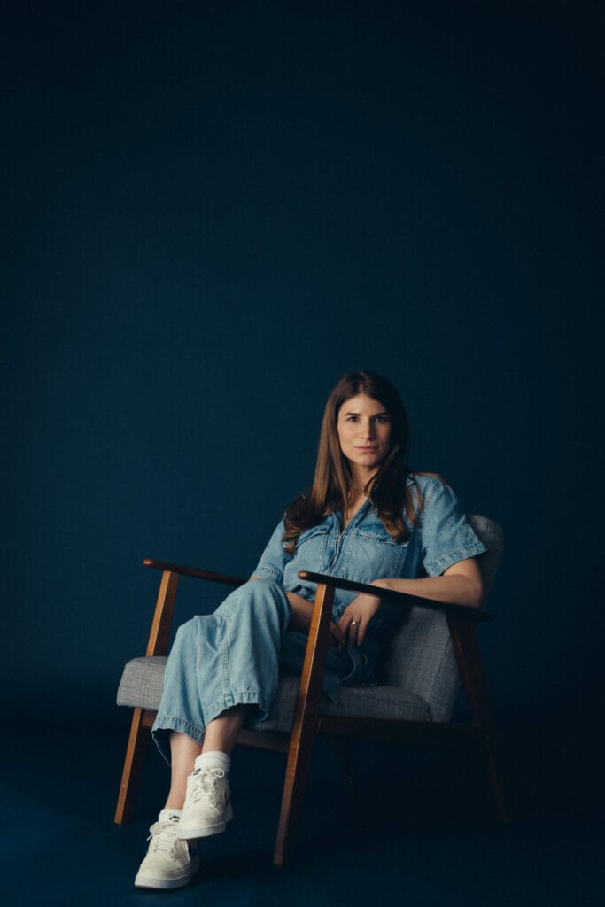 Vicky Purnell, with long brown hair, dressed in a light blue denim jumpsuit and white sneakers, sits in a mid-century modern armchair against a dark blue background, looking calmly at the camera.