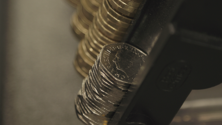 Close-up of stacks of coins, with a stack of dimes in focus, showing the profile of a person on the coin, held in a dark metal dispenser—reminiscent of a Royal Mint new launch featuring a UK icon.