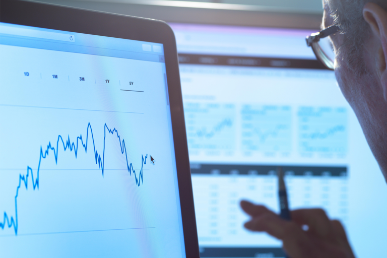 A person holding a pen analyzes financial data on computer screens, focusing on a fluctuating line graph and various charts—suggesting stock market, investment analysis, or monitoring venture capital dry powder allocations.