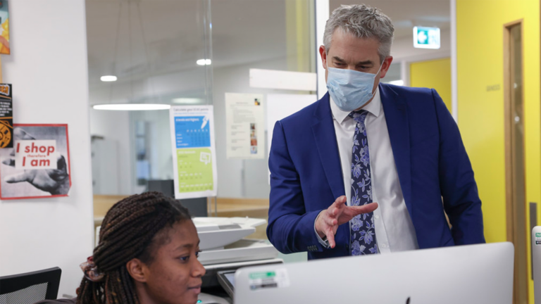 A man in a blue suit and face mask stands beside a seated woman at a computer, gesturing towards the screen as they discuss the pros and cons of a Major Conditions Strategy in an office with bright yellow walls.