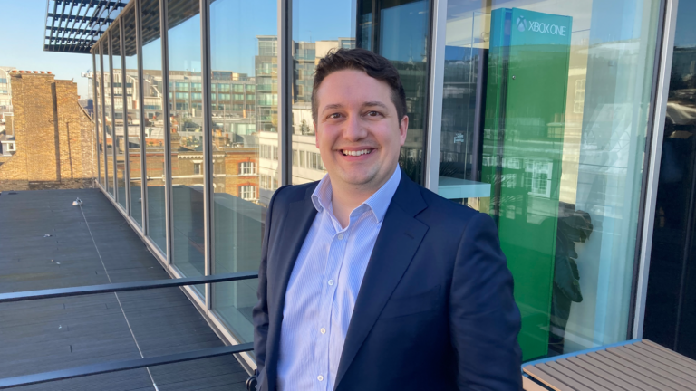 A man in a navy suit and light blue shirt smiles while standing on a rooftop terrace with glass walls, reflecting city buildings under a clear sky—a professional moment that captures the spirit of MHP Groups public affairs team.