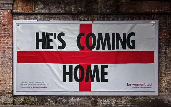 A billboard with a red St George’s Cross background reads “HE’S COMING HOME.” Below, Women’s Aid tackles football mischief and highlights domestic abuse, urging people to seek help at womensaid.org.uk.