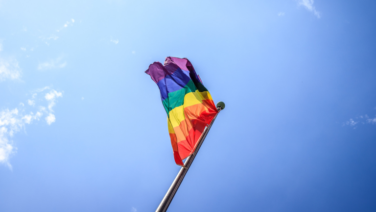 A rainbow-colored pride flag waves on a flagpole against a clear blue sky with a few small clouds, symbolizing year-round advocacy for the LGBTQ+ community.
