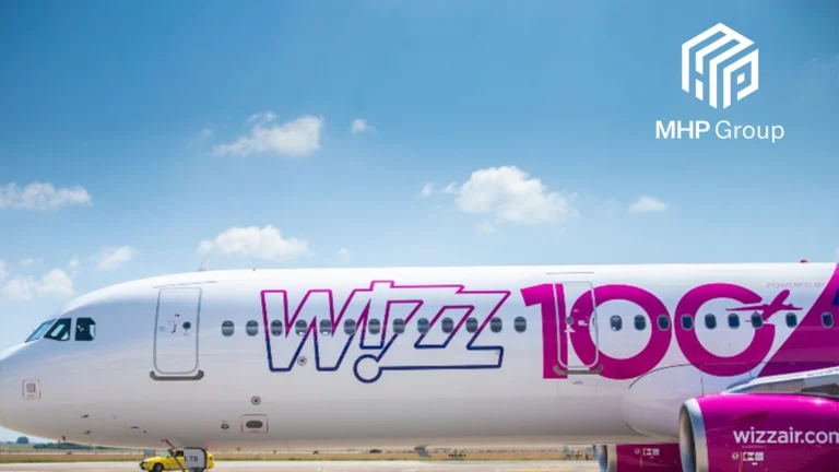 A Wizz Air airplane with 100 and wizzair.com on its side is parked on a runway. Showcasing MHP Groups expertise in financial communications, their logo appears in the upper right corner against a blue sky with scattered clouds.