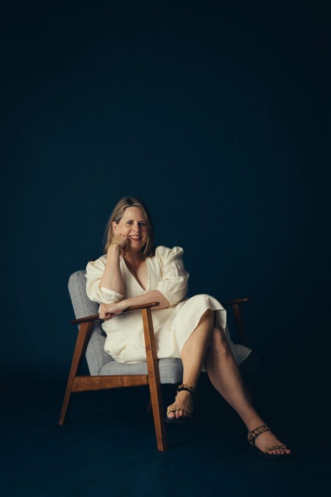 Chloe August, in a white dress, sits barefoot in a wooden armchair with a gray cushion, smiling softly and resting her head on one hand against a dark, solid-colored background.