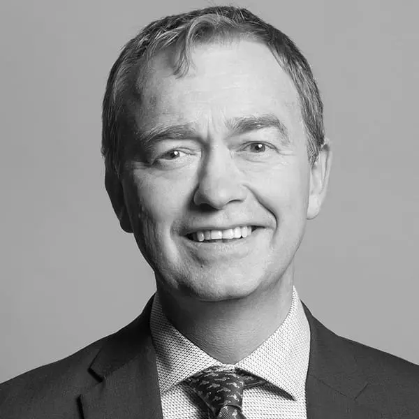 A middle-aged man in a suit and tie smiles at the camera against a plain background in a black and white portrait, evoking the professional demeanor of a UK Parliament MP.
