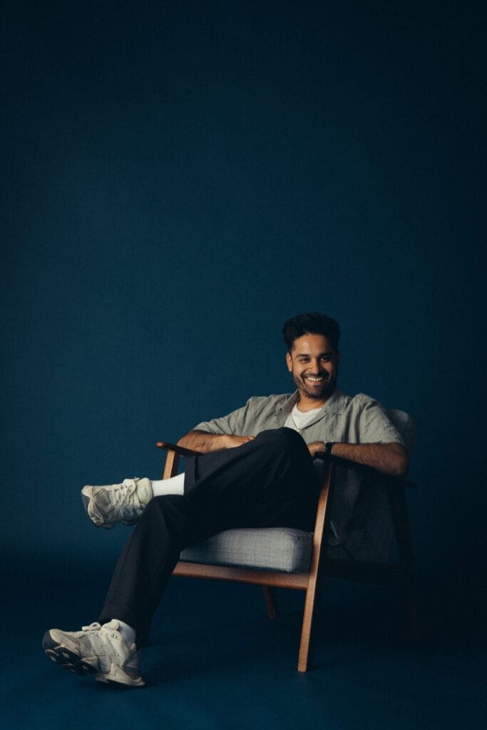 A man with dark hair and a beard smiles while sitting casually in a light gray armchair. He wears a light shirt, dark pants, and white sneakers, against a dark blue background.