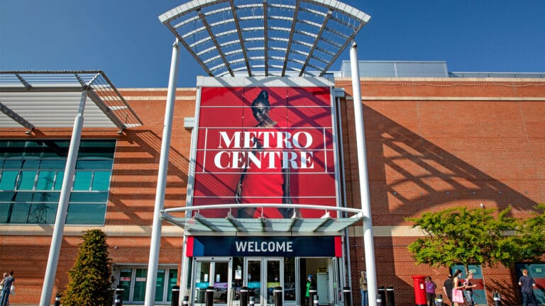 The entrance to Metrocentre shopping mall, managed by MHP Group, features a large red sign with METRO CENTRE in white letters, glass doors labeled WELCOME, and a modern metal canopy overhead.