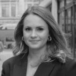 Gemma Lingham, with wavy hair and hoop earrings, smiles slightly while standing outdoors in a city setting. She is wearing a blazer, and the image is in black and white.