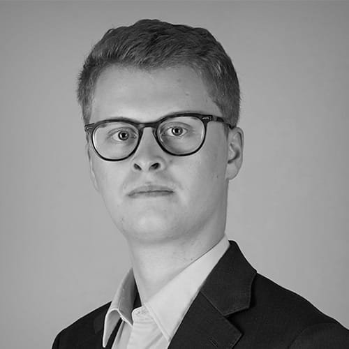 Matt Village, a young man with short light hair, glasses, a suit jacket, and a light-colored collared shirt, looks directly at the camera against a plain, light background. The black and white image highlights his poised presence.