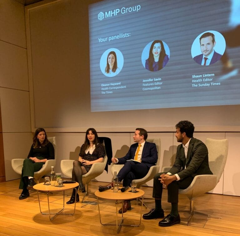 Four panelists sit on stage in modern chairs during a discussion event. Behind them, a screen displays the MHP Group logo and their names and titles, highlighting leading health journalists sharing fresh health insights, with water bottles on a small table in front.