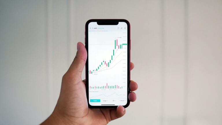 A hand holding a smartphone displays a financial trading app with a rising candlestick chart and technical indicators, set against a plain light background—highlighting how Money Matters in today’s fast-paced Financial Services world.