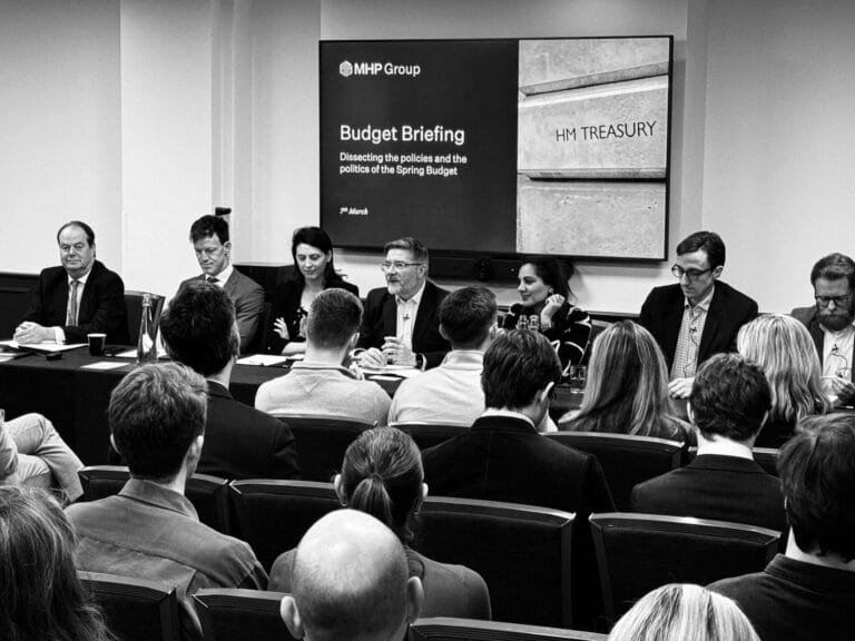 A panel of speakers from MHP sits at a long table addressing an audience during a Budget Briefing event. A screen behind them displays “MHP Group Budget Briefing” and “HM Treasury,” while attendees listen intently for key takeaways.