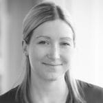 Black and white portrait of a woman with straight, light-colored hair, parted in the middle. She is smiling softly and looking into the camera, wearing a simple top. The background is softly blurred.