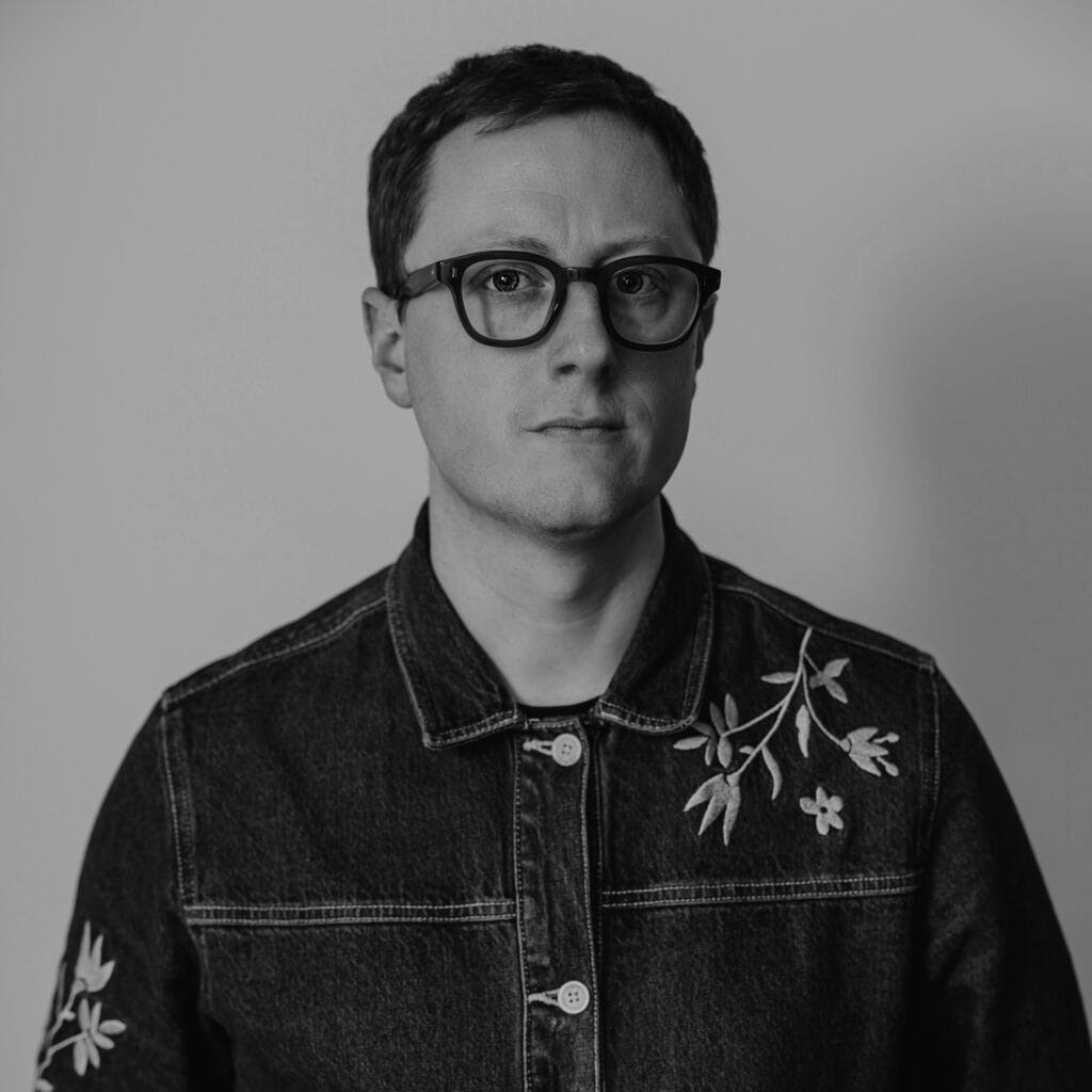 Jack Bedford, with short hair and glasses, wears a denim jacket adorned with embroidered floral designs, standing against a plain background in this black and white image.