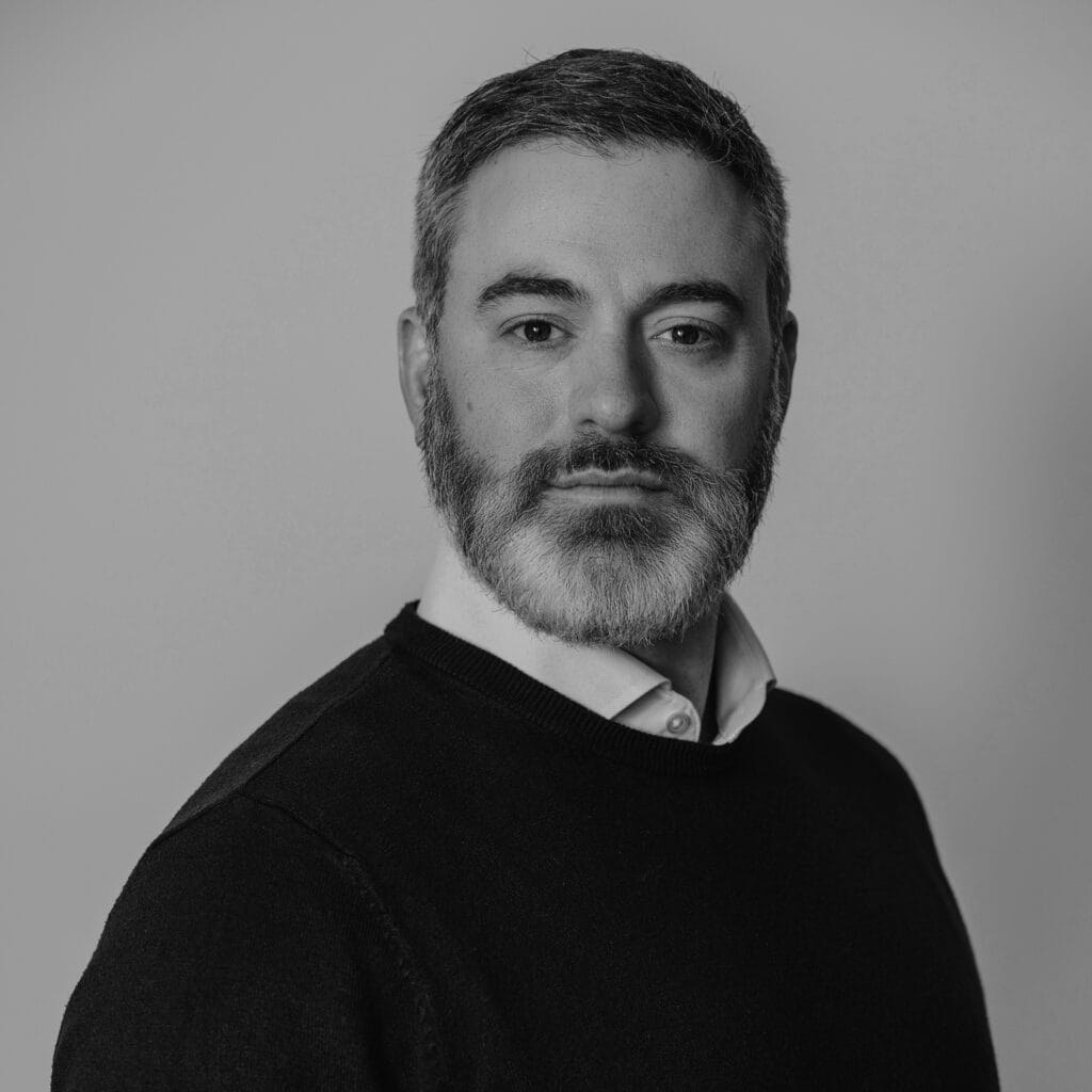 Black and white portrait of Matthew Francis, a man with short, dark hair and a beard, wearing a dark sweater over a collared shirt, looking confidently at the camera against a plain background.
