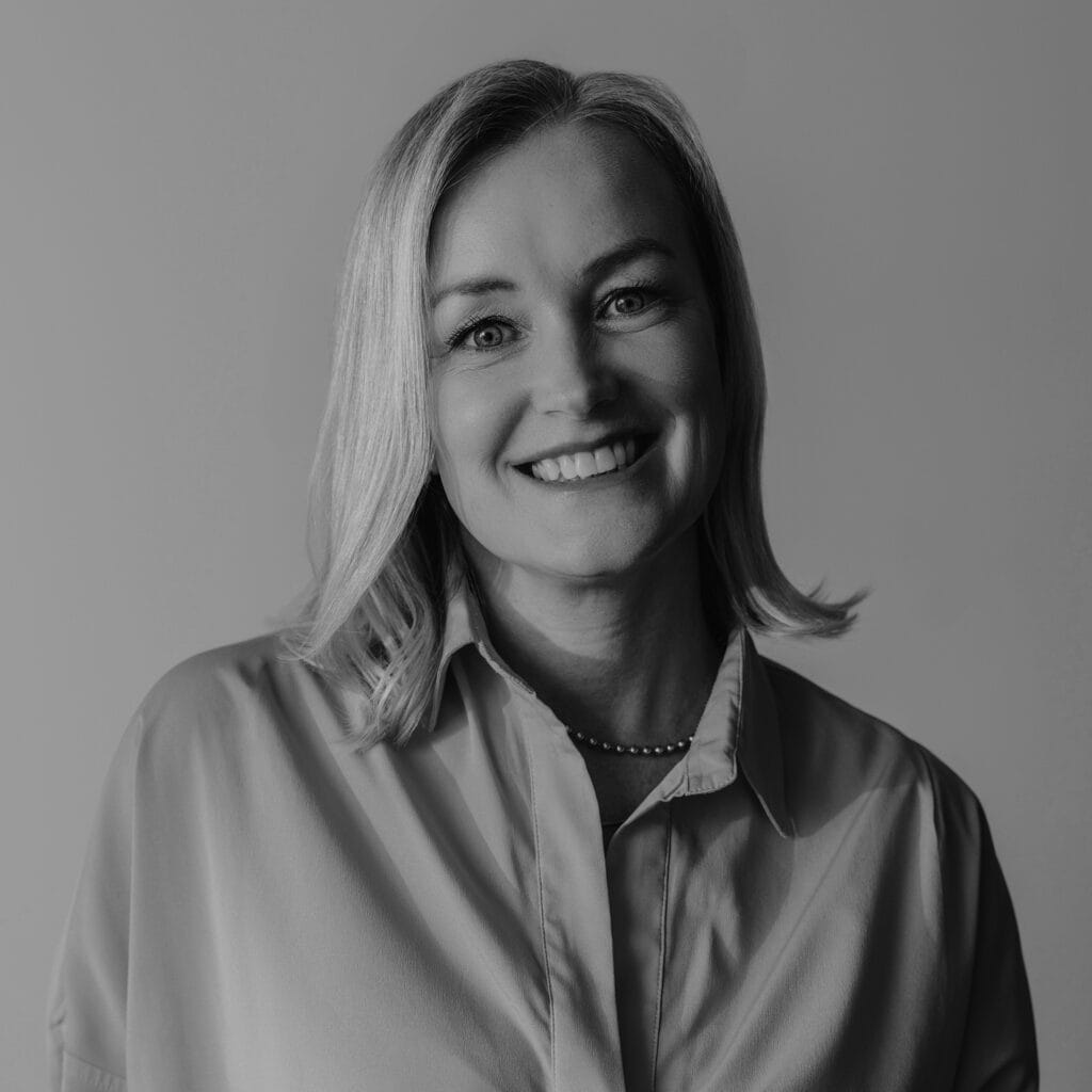A black-and-white portrait of Symone Jermin, a woman with shoulder-length light hair, wearing a collared shirt and a necklace, smiling at the camera against a plain background.