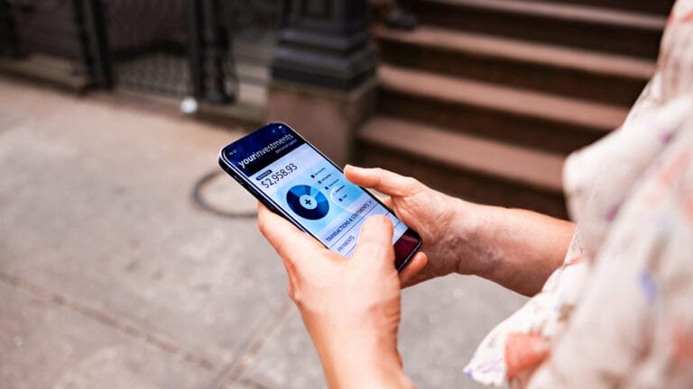 A person holding a smartphone on a city sidewalk uses the NextWealth app, checking their account balance and circular graph for smart financial advice.
