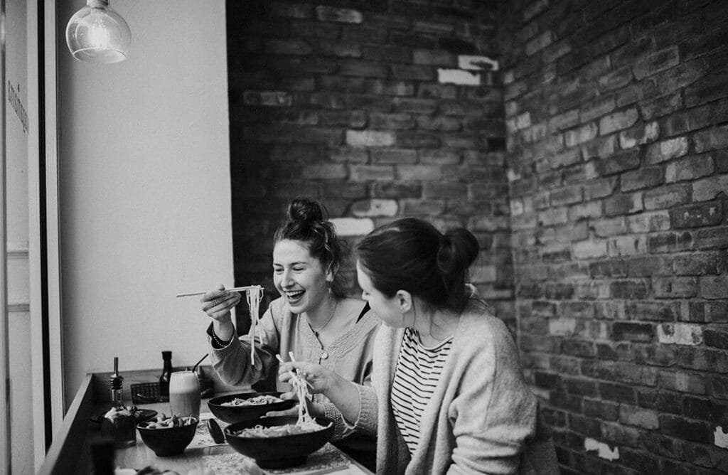 Two women sit at a table by a window, laughing and eating noodles with chopsticks in a cozy Restaurant Group venue with brick walls. Bowls of food and drinks are on the table, creating a relaxed and friendly atmosphere. MHP Group discuss supporting the move from AIM to FTSE 250.